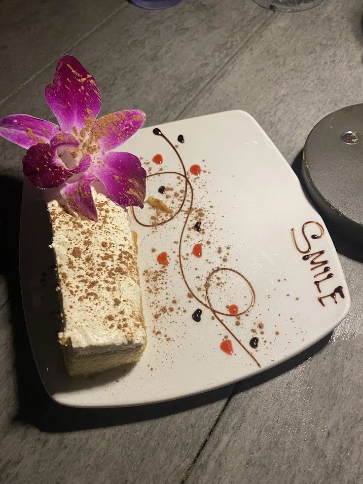 A slice of cheesecake on a white plate decorated with chocolate writing that says "SMILE" and various dots of chocolate and red sauce, garnished with a pink orchid flower.