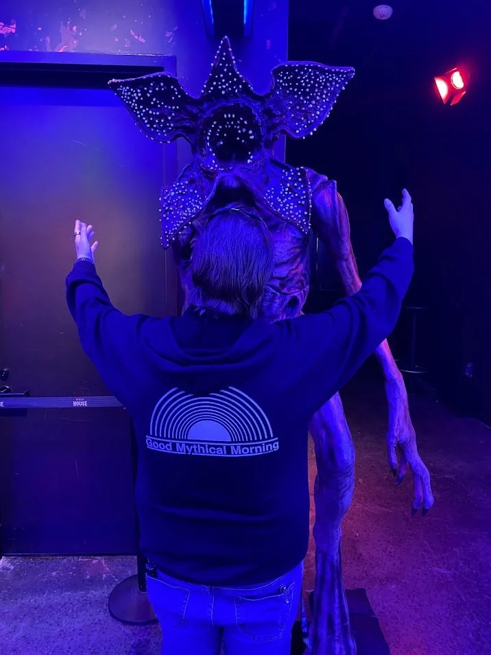 Person with long hair and a sweatshirt that says 'Good Mythical Morning' faces a large, alien-like creature with a head shaped like an octopus and decorated with glowing dots, in a dark, neon-lit room.