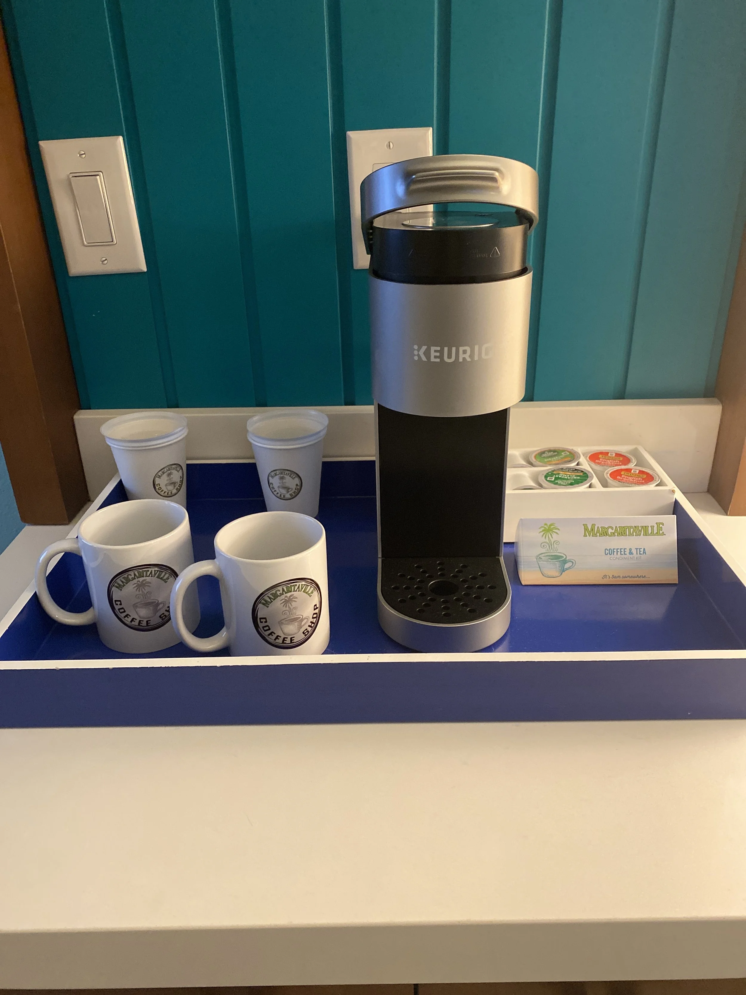 A coffee station with a Keurig coffee maker, two white mugs and two white cups, along with a tray of coffee flavor packets and a small sign reading 'Margaritaville Coffee & Tea' against a teal wall.