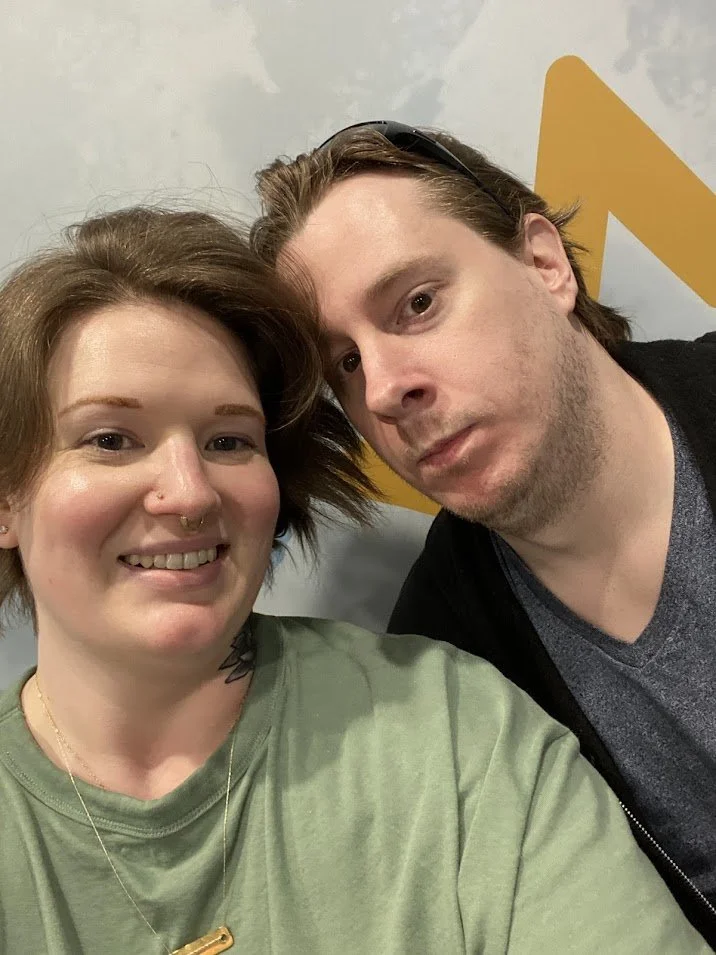 A woman with short red hair and a septum piercing smiling next to a man with brown hair and a slight beard, both taking a selfie indoors.