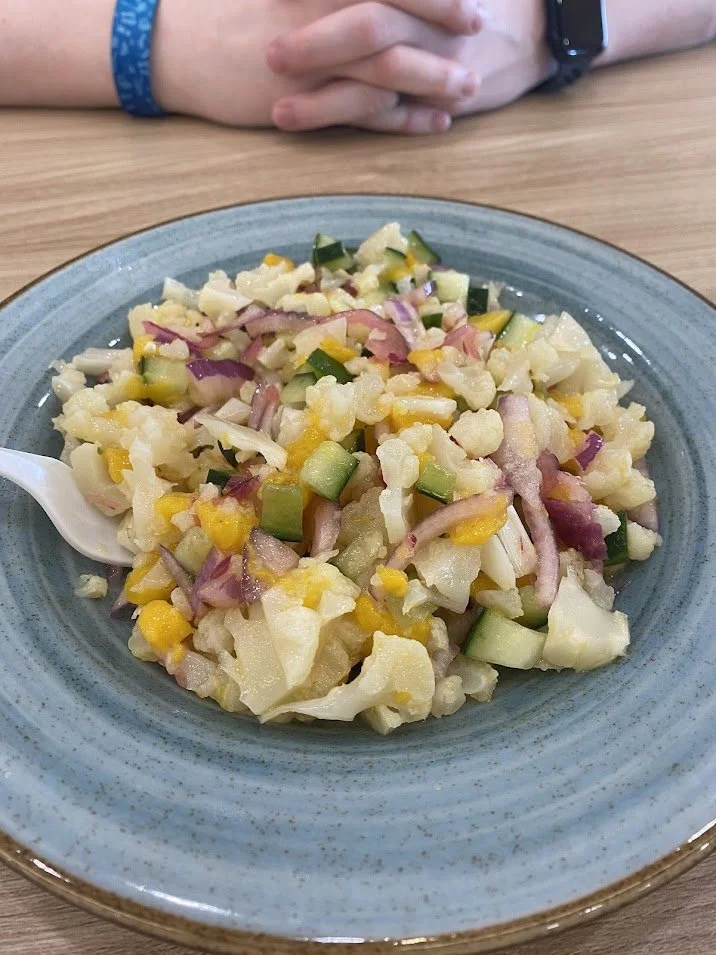 A plate of potato salad with chopped onions, celery, and pickles on a wooden table with a person's hands clasped in the background.