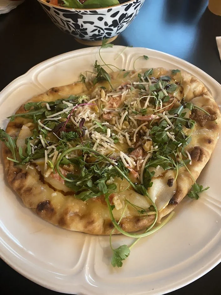 A thin-crust pizza topped with cheese, shredded cheese, mixed greens, and sprouts on a white plate, with a bowl of salad in the background.