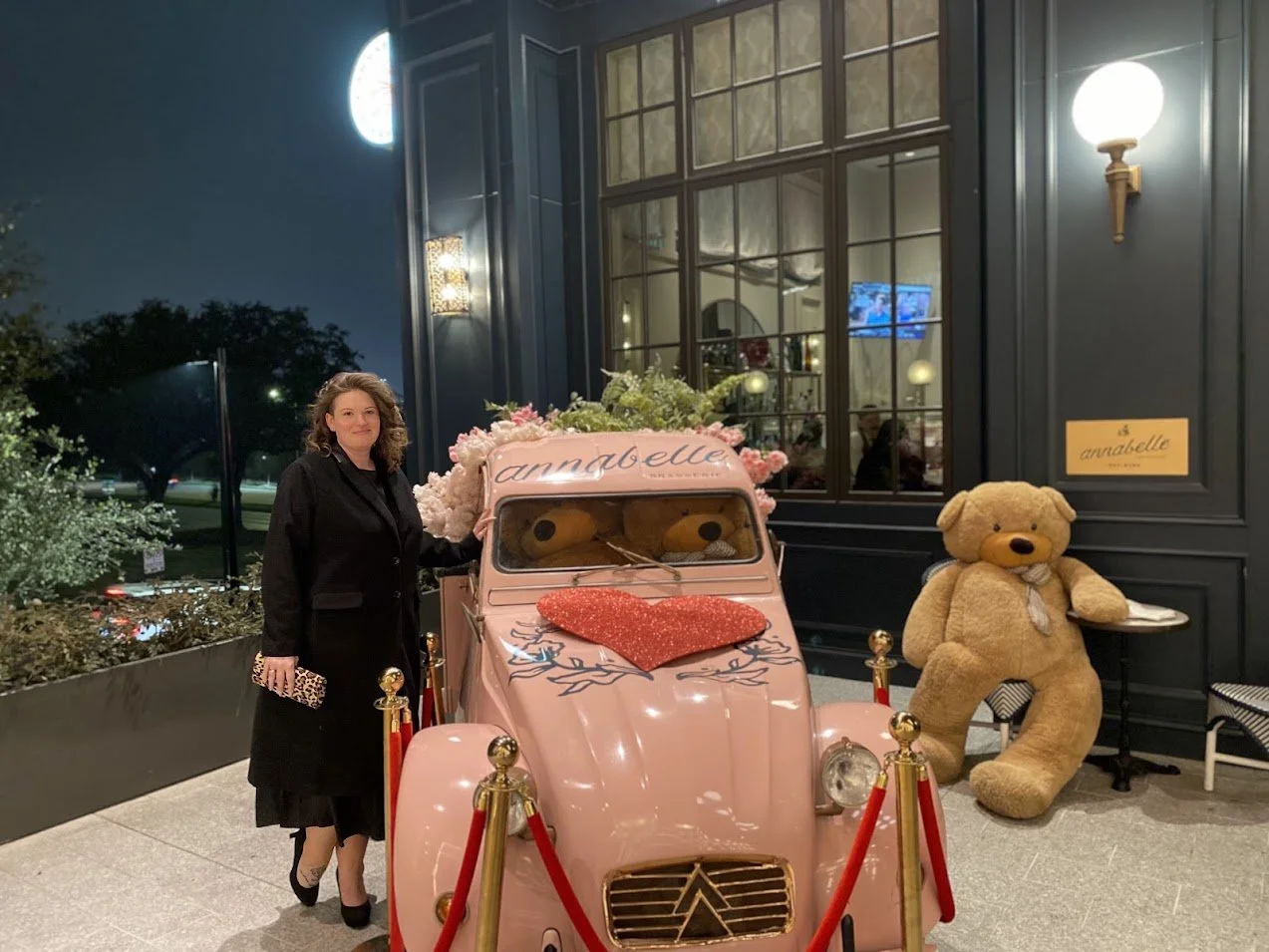 A woman in a black coat standing next to a pink vintage car decorated with flowers and a large red heart on the hood, outside at night in front of a dark building with large windows. There is a large teddy bear sitting on a small table to the right o
