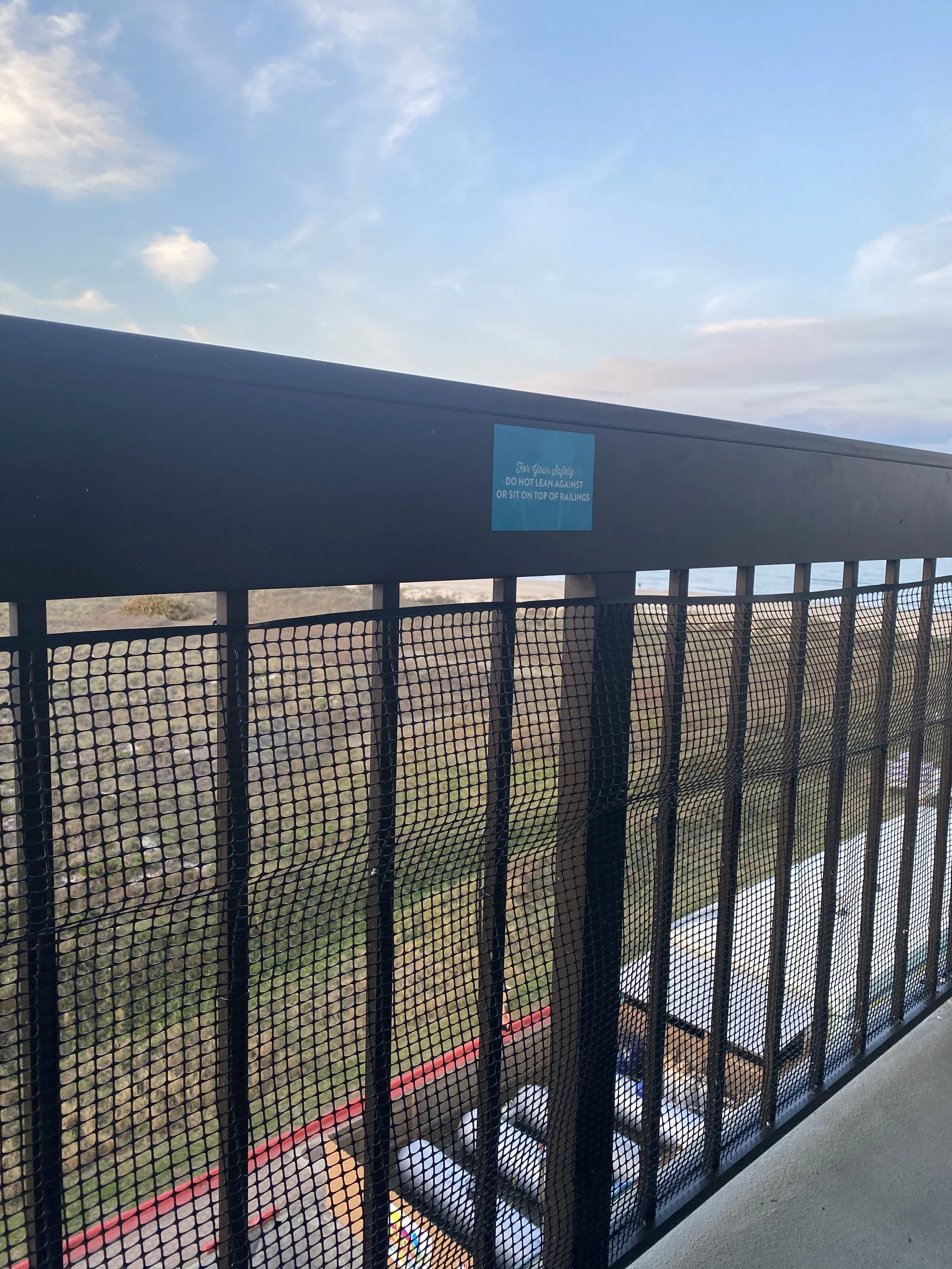 View from a balcony showing a black railing with a sign that reads 'For Your Safety, Do Not Lean Against or Sit on Top of Railings.' The sky is partly cloudy with a few clouds and the landscape below includes trees, buildings, and a parking lot.