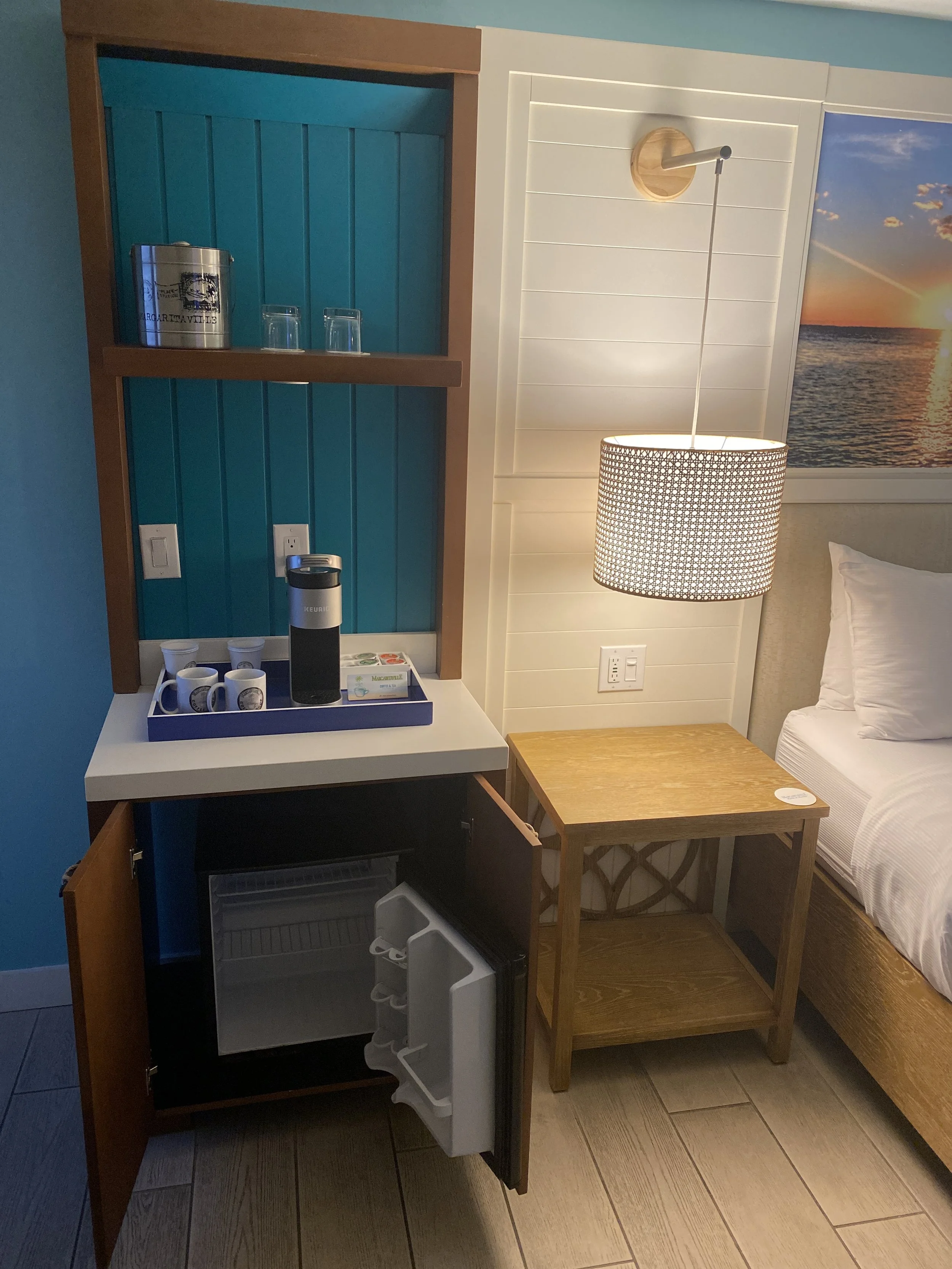 Room with a small refrigerator, a wooden nightstand, a hanging lampshade, a bed, and a wall art of a sunset over the water.