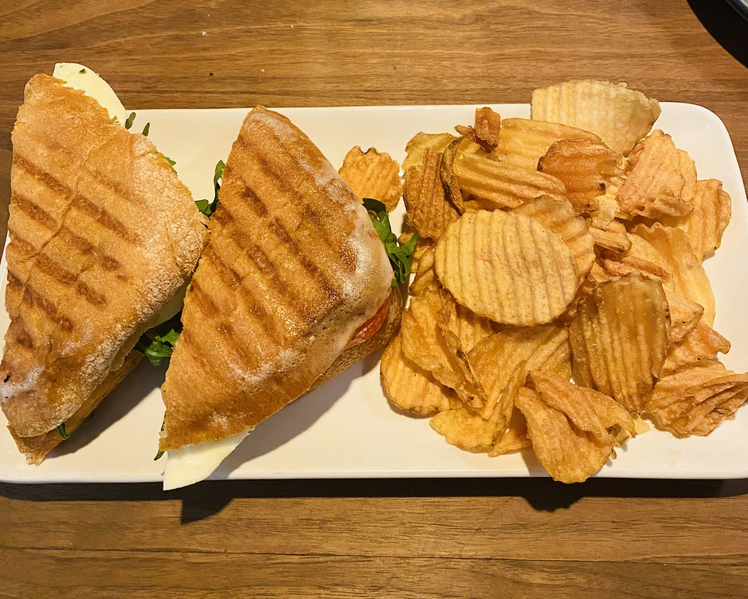 A grilled sandwich on a white rectangular plate with crinkle-cut potato chips on the side, placed on a wooden table.