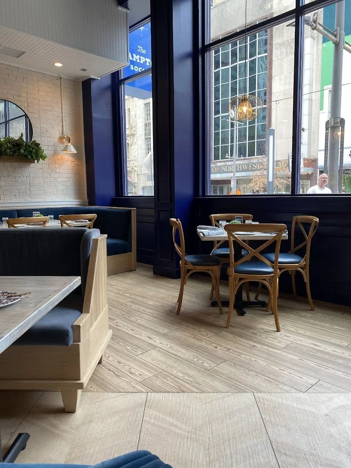 Interior of a modern restaurant with wooden and upholstered seating, large windows, and city buildings visible outside.