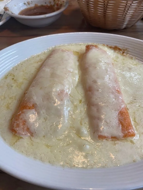 Two cheese enchiladas covered in a creamy white sauce on a white plate with a small bowl of sauce and a basket in the background.