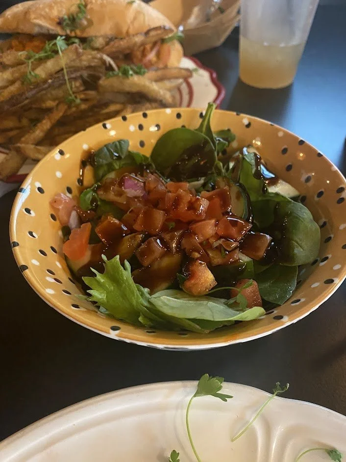 A bowl of fresh mixed greens with diced tomatoes, red onions, and a drizzle of balsamic vinaigrette. There is a plate of fried chicken wings and a sandwich in the background, along with a glass of iced beverage on a dark table.
