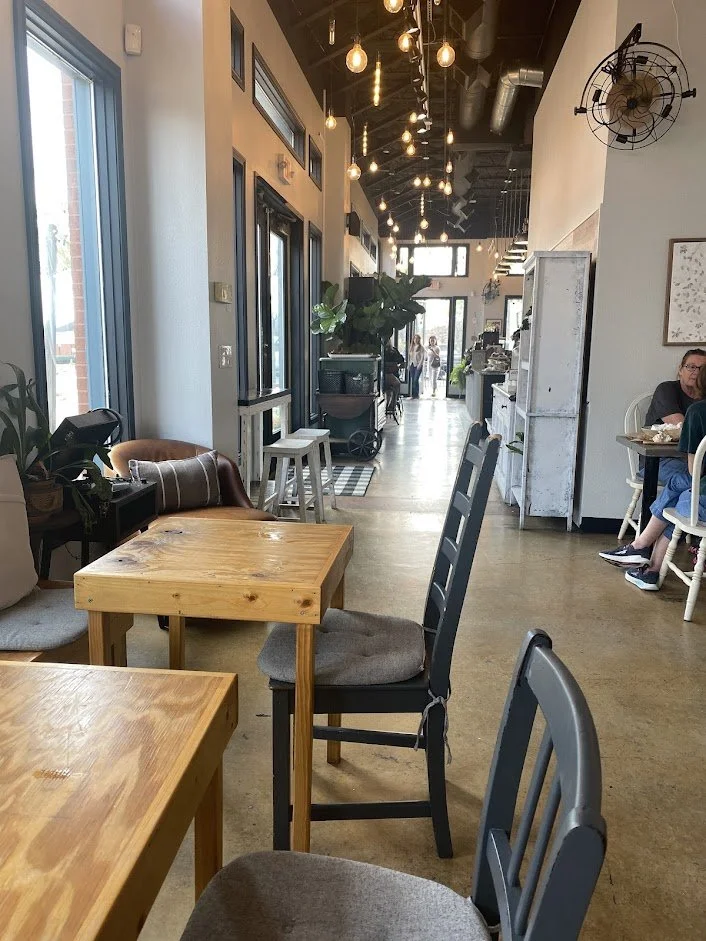 Interior of a cozy cafe with wooden tables, black chairs with gray cushions, hanging Edison bulb lights, large windows on the left, potted plants, a woman sitting at a table on the right, and a group of people walking towards the exit at the back.