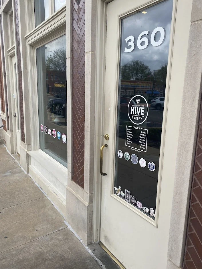  storefront with large glass doors and windows. The door has a sign indicating the address number "360" and the name "Hive Bakery" with business hours. There are various stickers and decals on the door and window, and the building has brick and stone