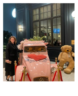 A woman standing next to a pink car decorated with a large pink bow and a sign reading 'Congratulations.' The car has a plush bear on the front seat and is surrounded by red rope barriers at night outside a building with large windows.