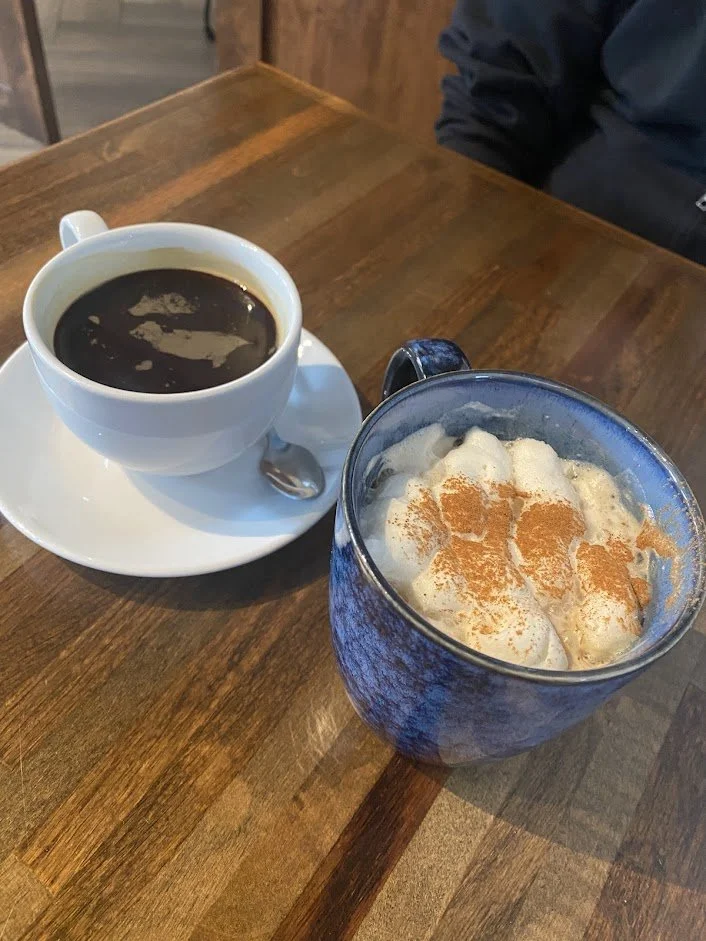 A white coffee cup filled with black coffee on a white saucer and a blue mug with white whipped cream topped with cinnamon, on a wooden table.