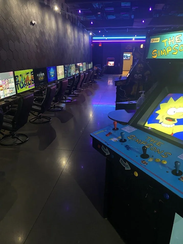 An arcade with a row of computer gaming stations on the left, and classic arcade games like The Simpsons on the right, under colorful neon lighting.