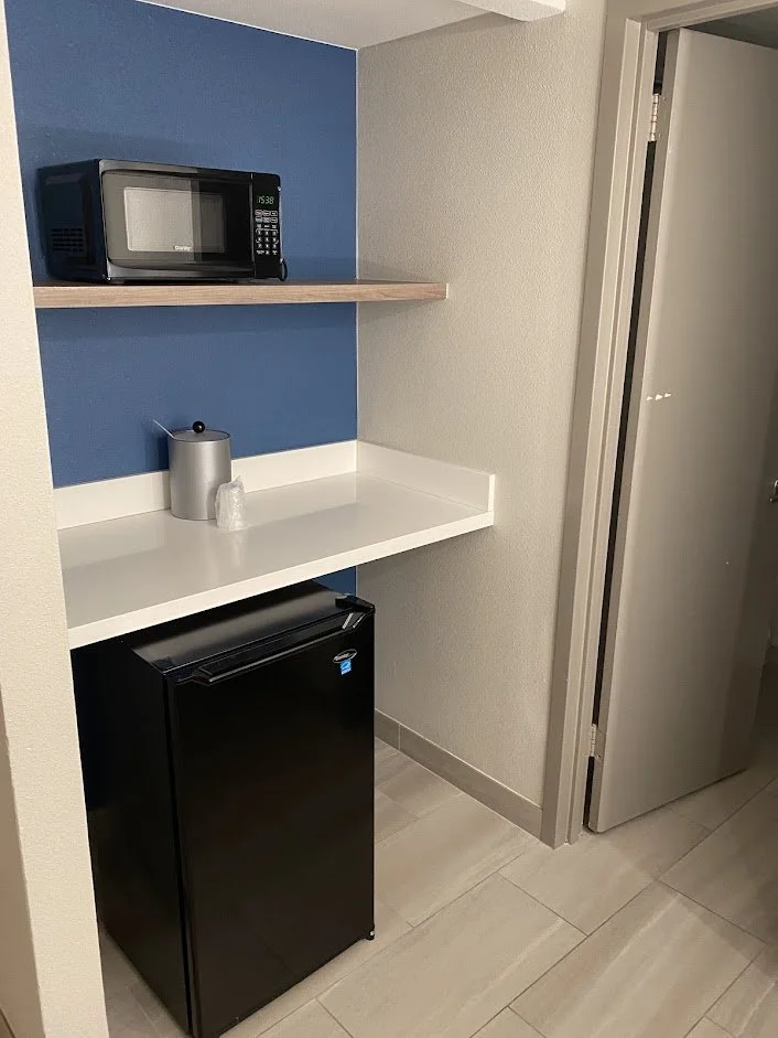 A small kitchenette with a black mini fridge, a white countertop with a silver container and a small item, a wooden shelf with a microwave, and a doorway to another room.