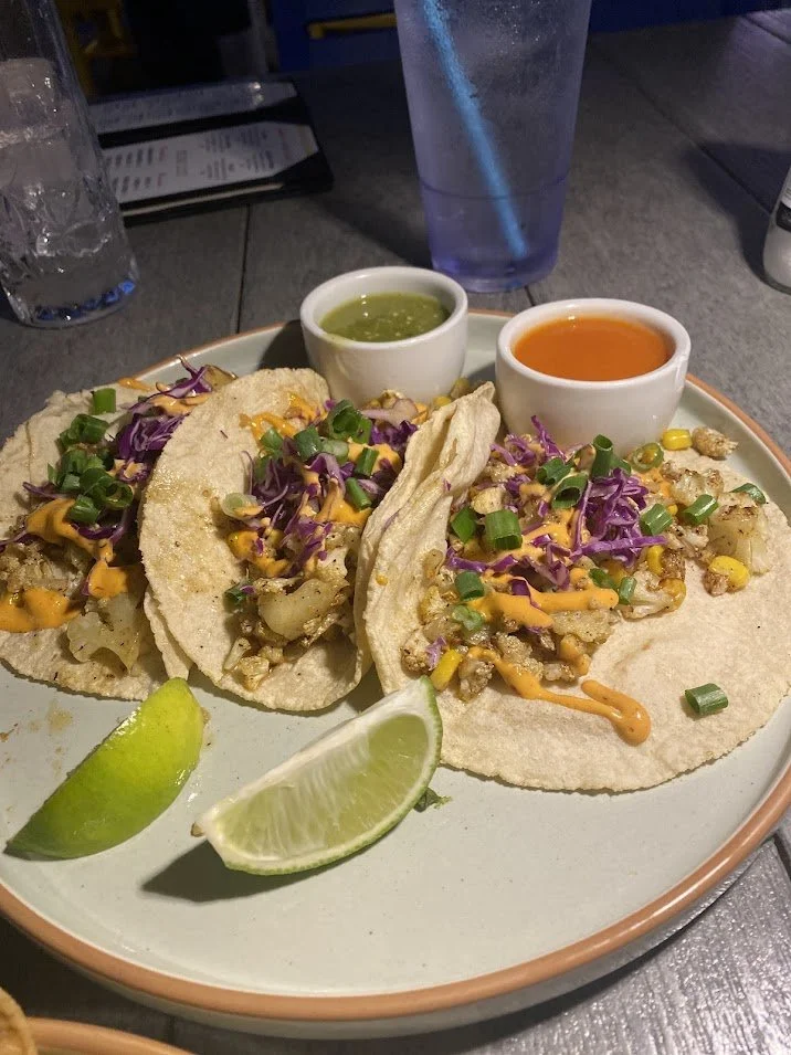 Three tacos with chopped vegetables, shredded cheese, sauces, and lime wedges on a plate, with drinks in the background.