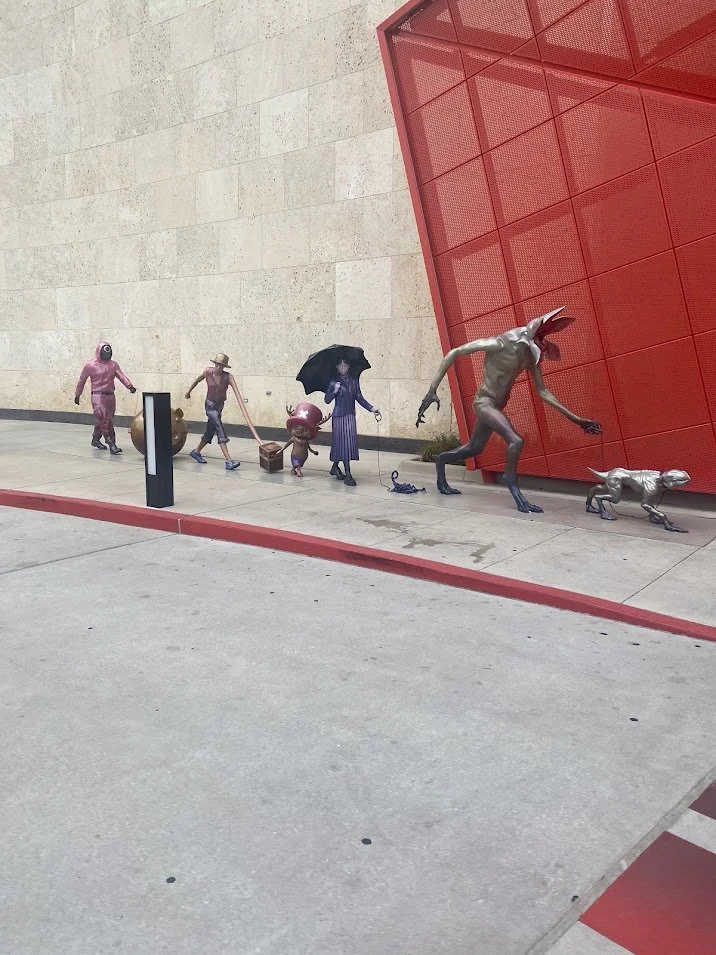 Art installation of whimsical, metallic sculptures of various fantasy creatures and characters, including a horned animal and a small dog, placed along a building wall with a red geometric structure.