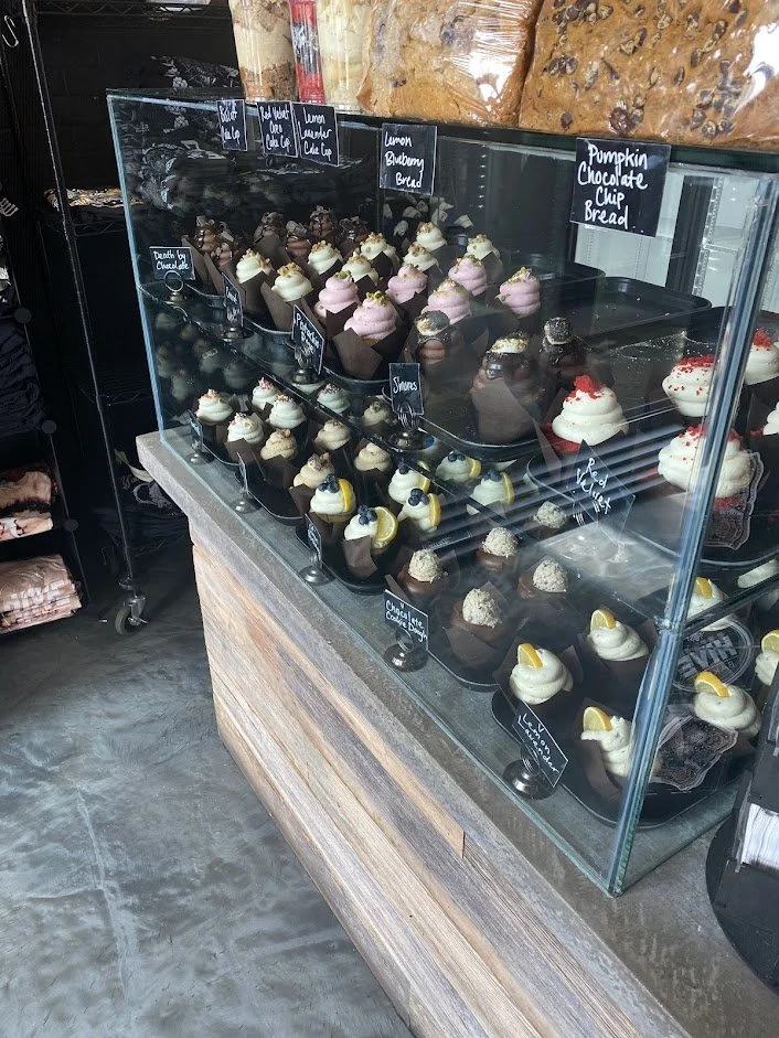 Display case with assorted cupcakes in a bakery, labeled with flavors such as Lemon Blueberry Bread, Pumpkin Chocolate Chip Bread, and Chocolate Stout.