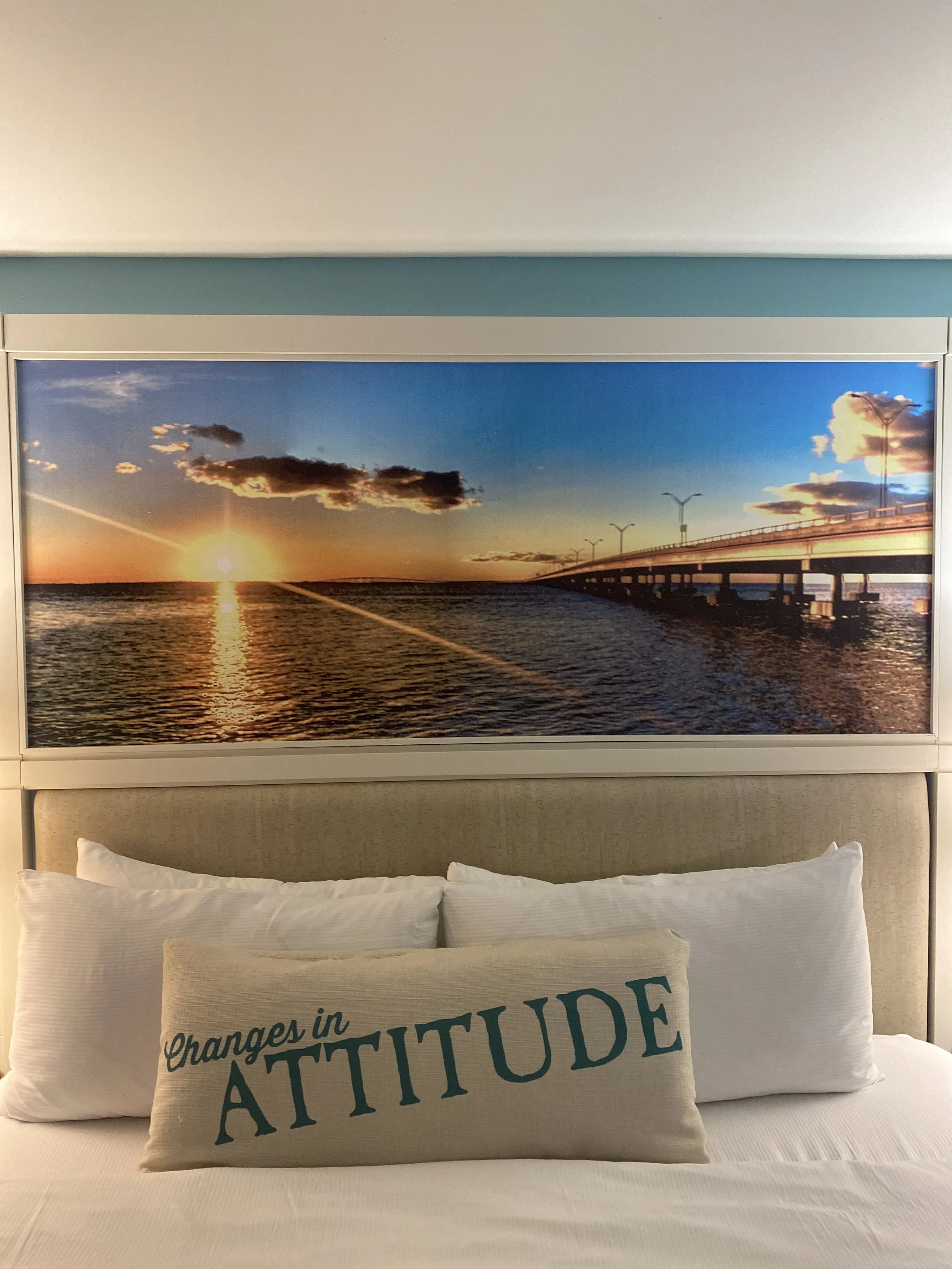 A bed with white pillows and a beige pillow that has the words "Changes in ATTITUDE" printed on it. Above the bed, there is a large framed picture of a sunset over a body of water with a bridge on the right side.