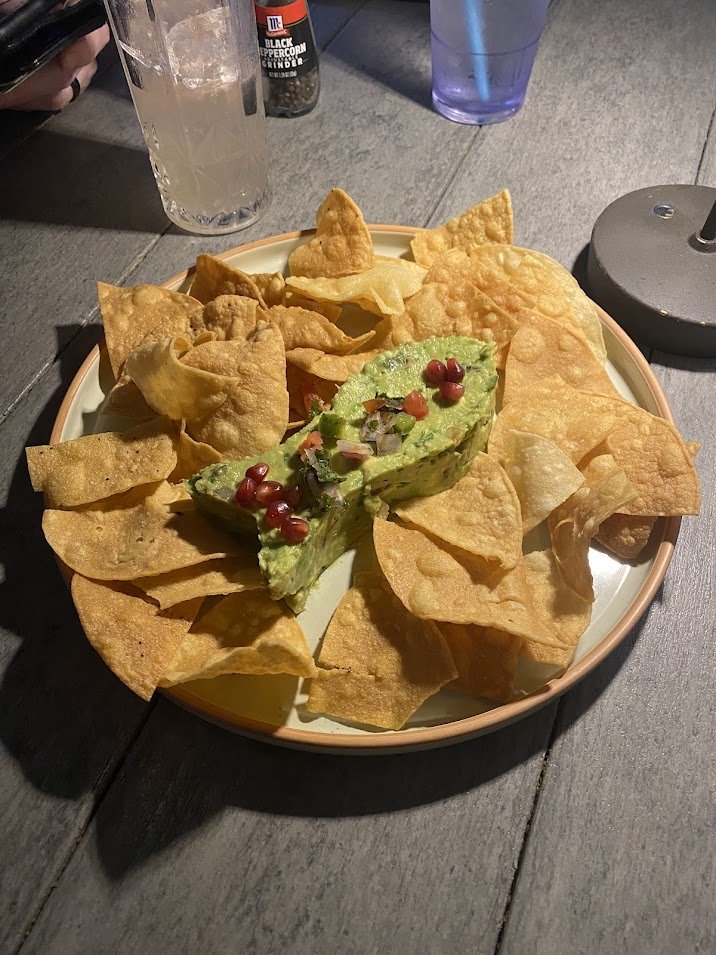 A plate of tortilla chips topped with guacamole garnished with pomegranate seeds and chopped onions, on a table with drinks and condiments.