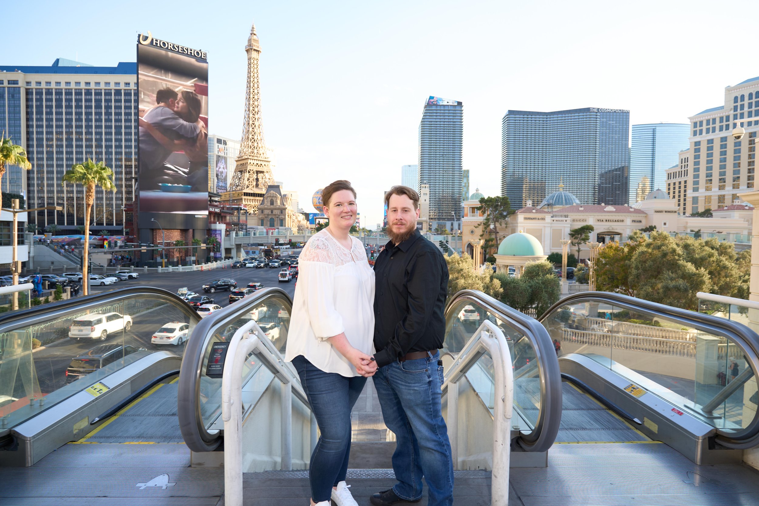 A couple holding hands on an escalator in front of the Las Vegas skyline, including the Eiffel Tower replica and various tall buildings.