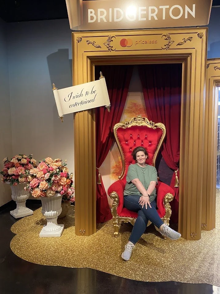 A woman sitting on a red and gold throne with a red curtain behind her. There are two large flower arrangements in white vases on the left. Above, a sign says "BRIDGERTON" and a smaller sign with the Mastercard logo and the words "price.ess" near the