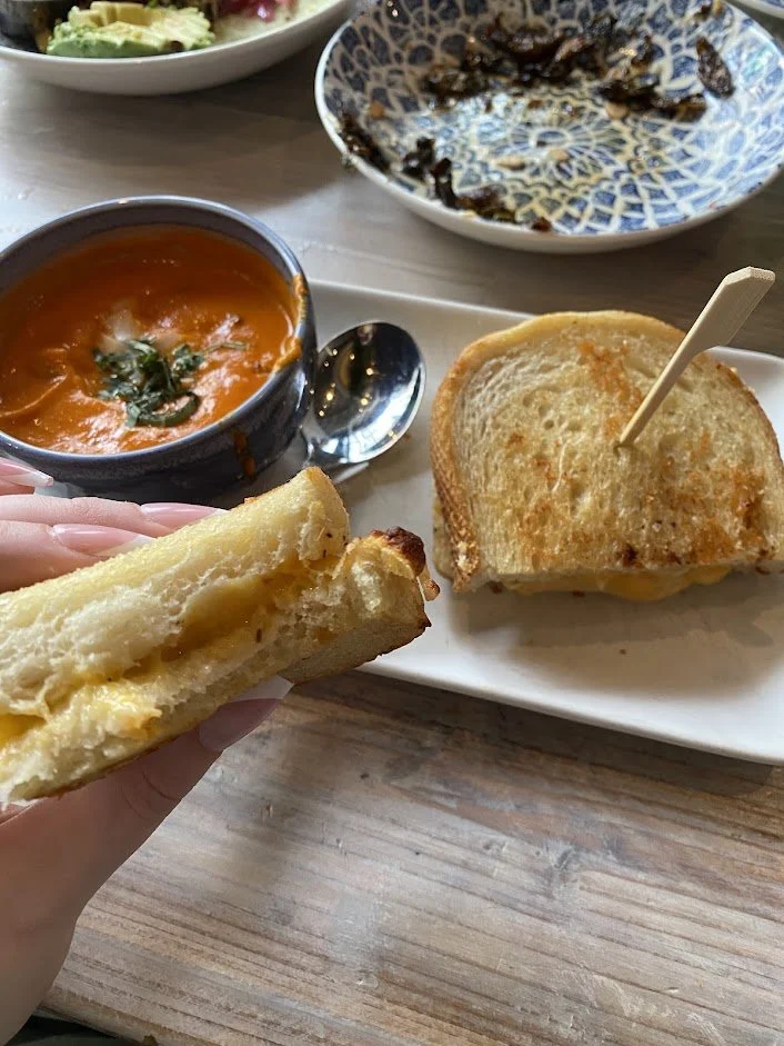 A hand holding a grilled cheese sandwich with melted cheese. On the table, there is a bowl of tomato soup topped with basil, a grilled cheese sandwich on a white plate, and a blue and white patterned bowl with some remnants of a dark dish. In the bac