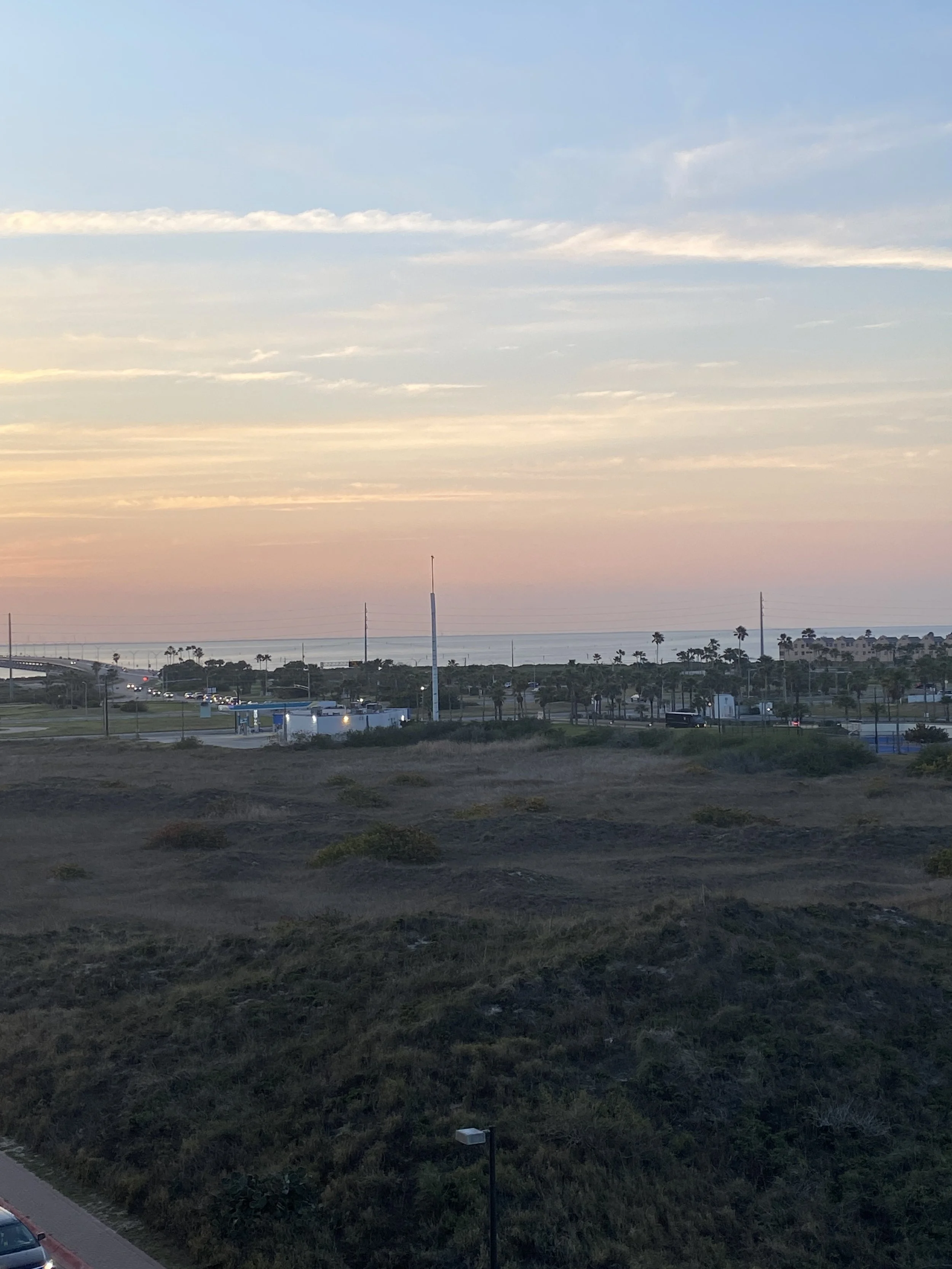 Sunset over a coastal landscape with a highway, palm trees, and a body of water in the background.