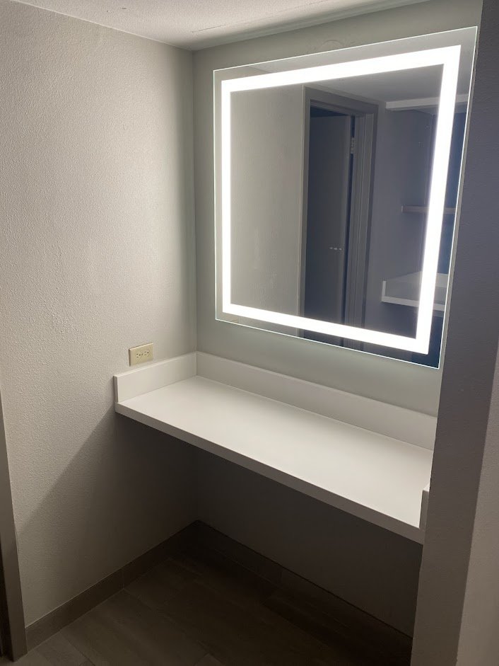 A brightly lit mirror with LED edges mounted on a wall above a white countertop in a bathroom or dressing area, with a doorway reflected in the mirror.
