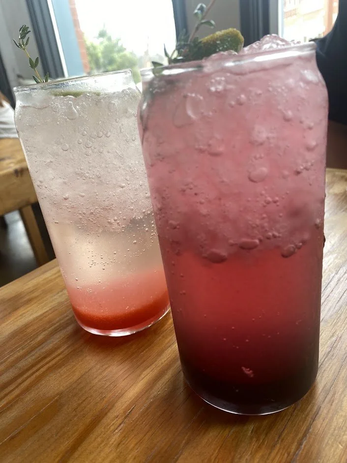 Two tall glasses of flavored soda with ice and fruit garnishes, one pink and one clear with a hint of pink, on a wooden table near a window.
