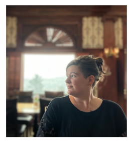 A woman with short dark hair, wearing a black sweater, looking to her right in a warmly lit restaurant or café with wooden interior and a large window in the background.