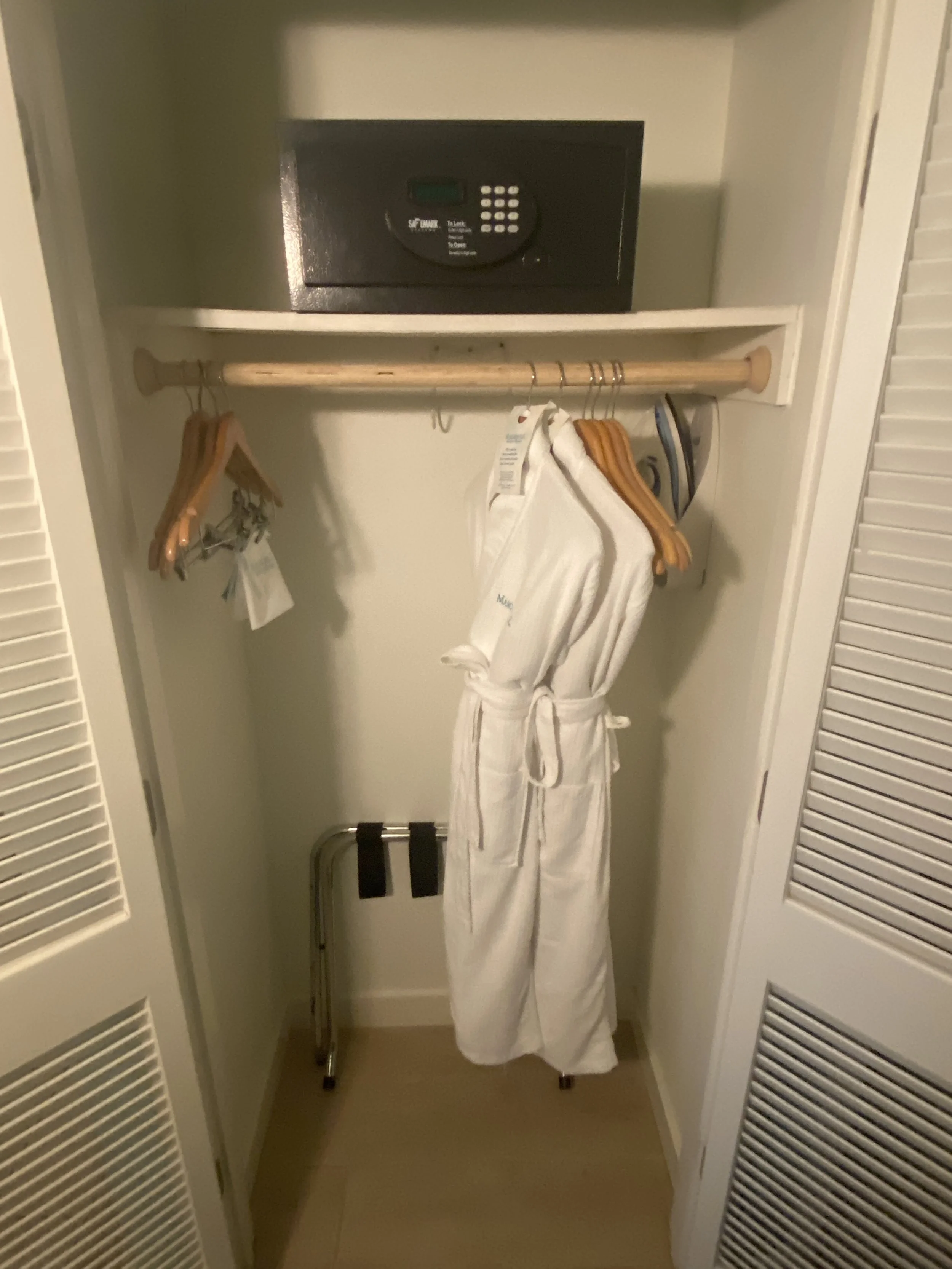 An empty hotel closet with a safe on the top shelf, wooden hangers on a rod, and a white bathrobe hanging in the middle.