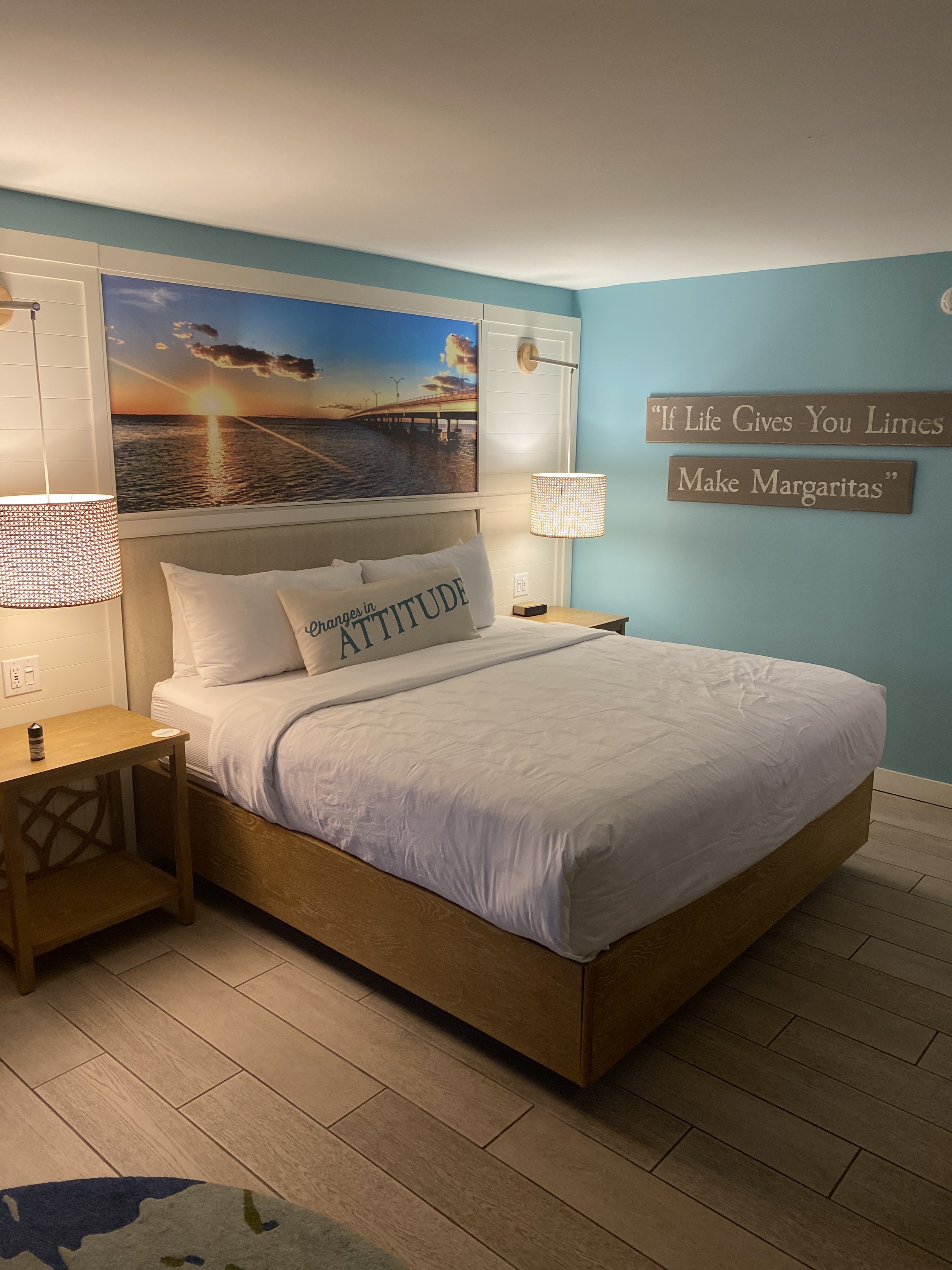 A bedroom with a king-sized bed, a large framed sunset or sunrise picture above the headboard, two nightstands with lamps, and a decorative pillow with the text ‘Changes in ATTITUDE’ on it. There are wall signs with the quote ‘If Life Gives You Lemon