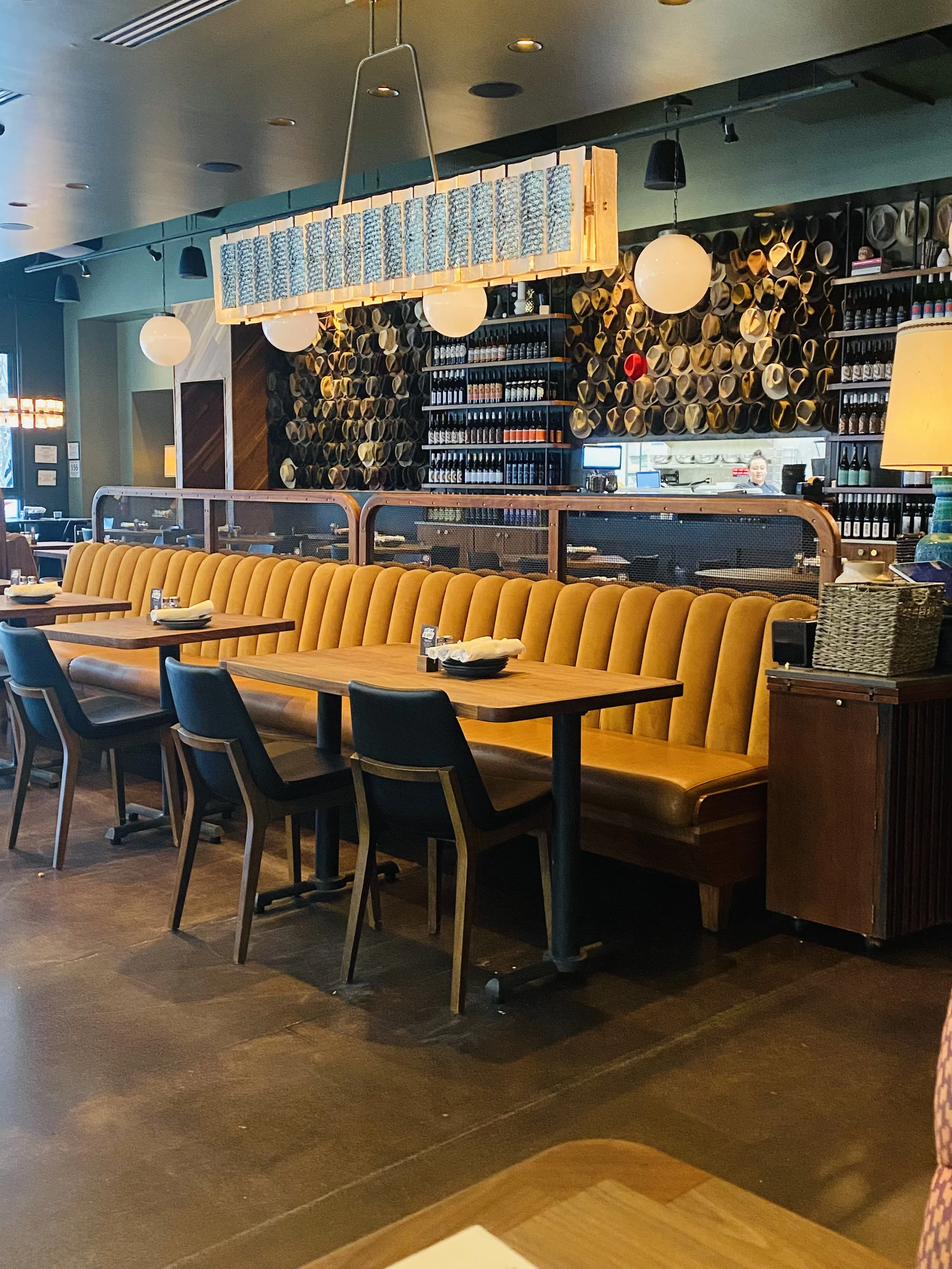 Empty booth with a mustard-colored upholstered bench, rectangular wooden tables, and dark chairs in a modern restaurant interior. The background features shelves with wine bottles and a wall with decorative wine corks. Overhead lighting includes a wo