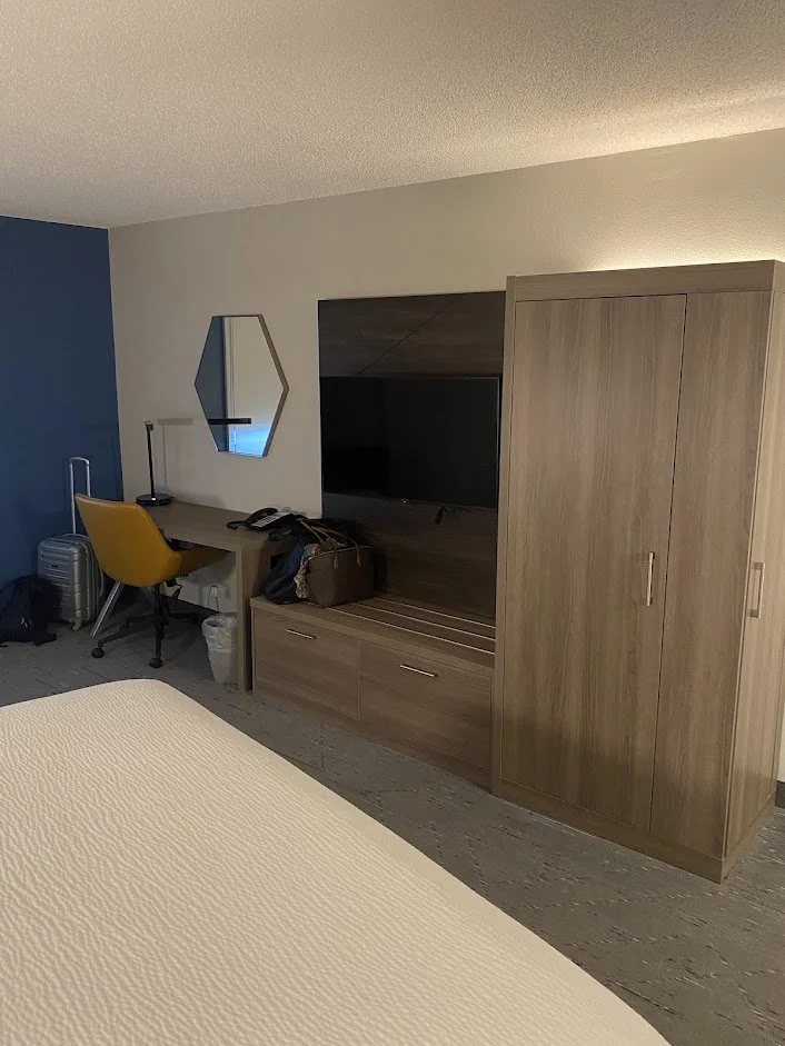 Hotel room with a bed, a wooden wardrobe, a TV in a wooden cabinet, a desk with a yellow chair, a mirror, a luggage, and some personal items.