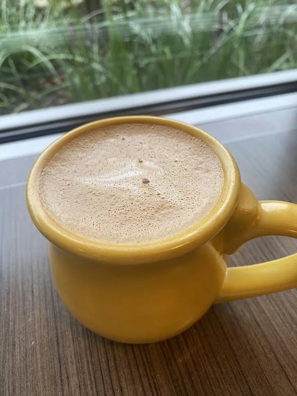 Yellow mug filled with frothy coffee on a wooden table near a window showing greenery outside.