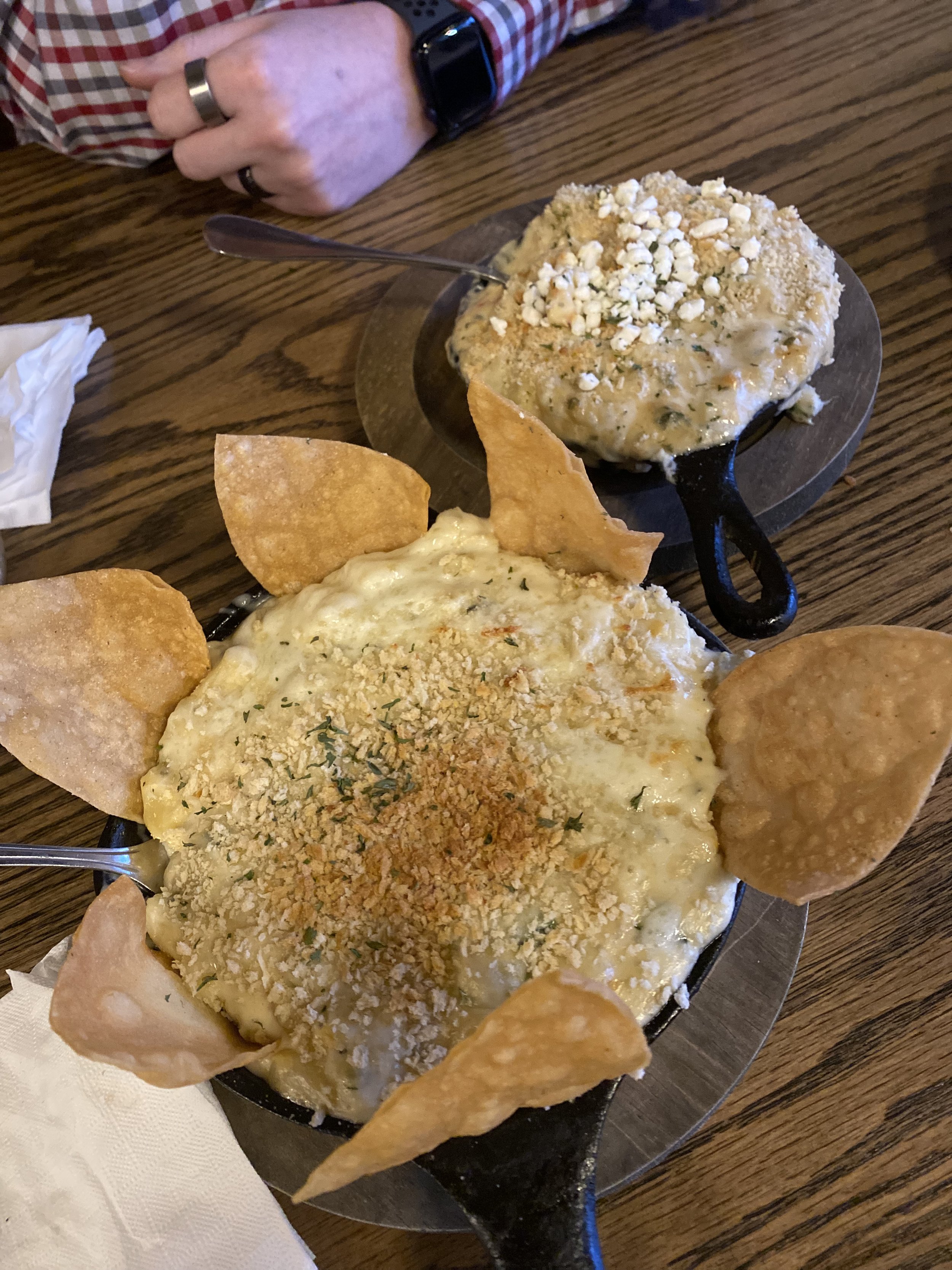 Two sizzler plates with cheesy baked dip topped with crushed crackers and crunchy tortilla chips, on a wooden table.