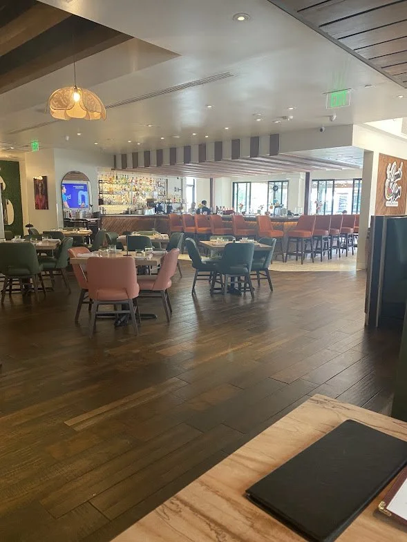 Interior of a restaurant with wooden flooring, colorful cushioned chairs around tables, a bar area in the background, and large windows letting in natural light.