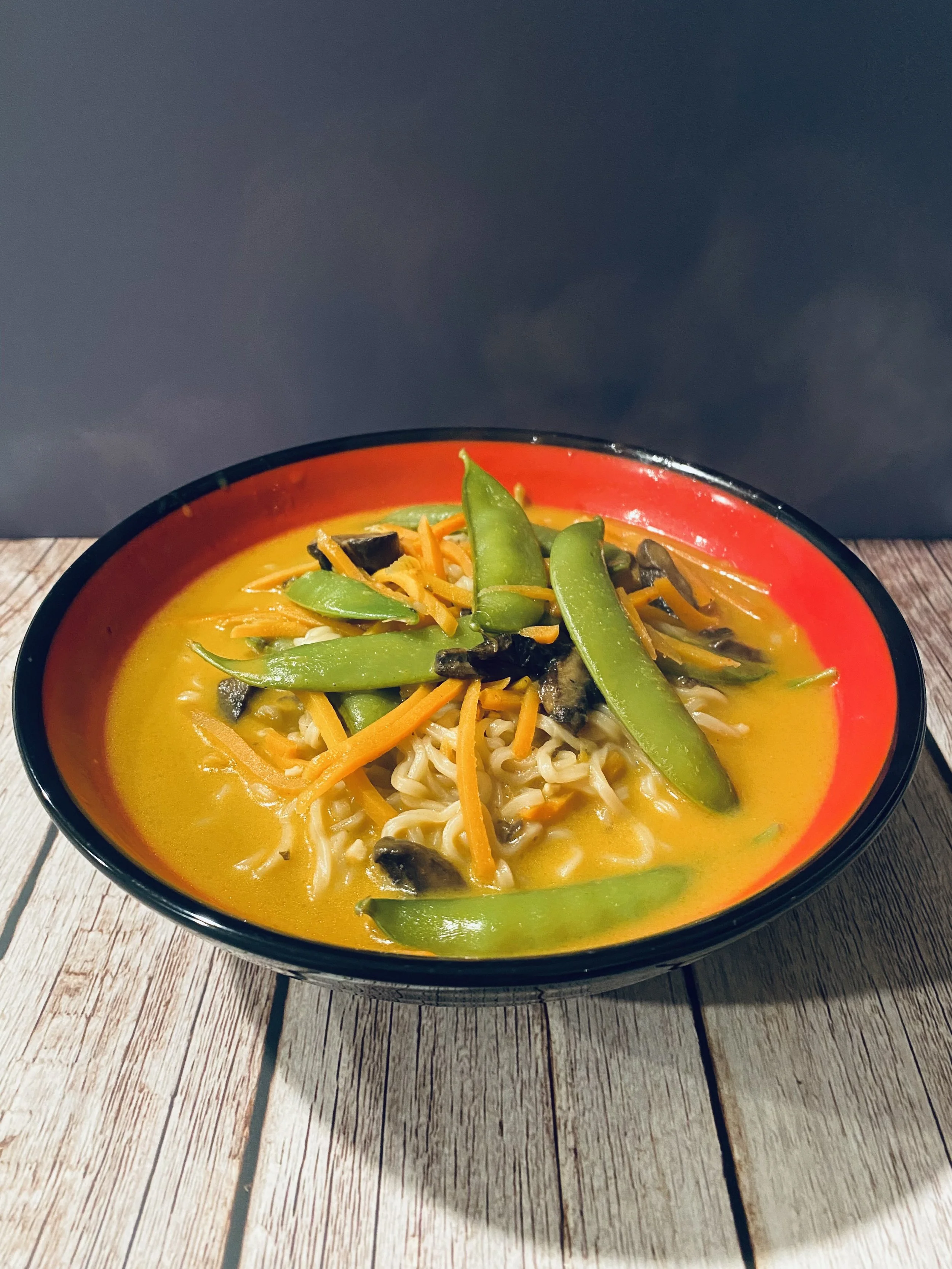 A bowl of ramen with yellow broth, noodles, and topped with snow peas, shredded carrots, and black mushrooms on a wooden surface.