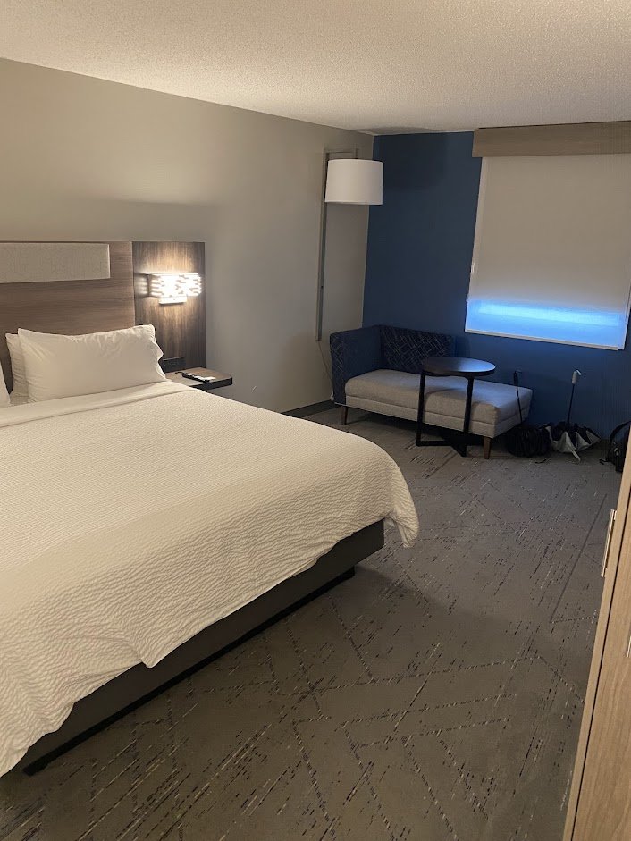 A hotel room with a large bed, white bedding, a nightstand with a phone, a wall-mounted lamp, a blue accent wall, a window with a closed blind, a small sitting area with a chair, a side table, and a floor lamp.