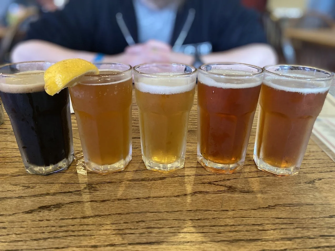 Five shot glasses of beer arranged in a row on a wooden table, with the first glass containing a dark stout garnished with a lemon wedge, and the remaining four glasses filled with different types of beer in varying shades from pale to amber.