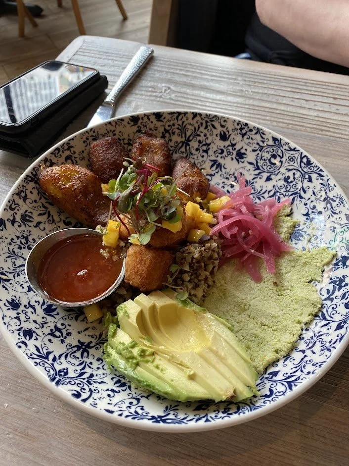 A decorative bowl containing three fried potatoes, avocado slices, pickled onions, a small cup of salsa, garnished with microgreens, served with yellow rice, black beans, and diced pineapple.