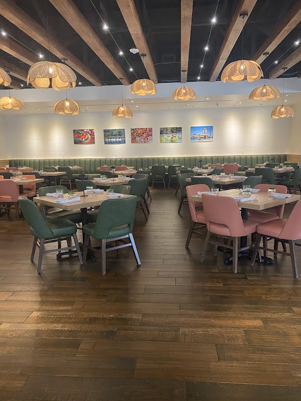 An empty restaurant with a wooden floor, multiple tables with pink and green chairs, ceiling lights, and colorful paintings on the wall.