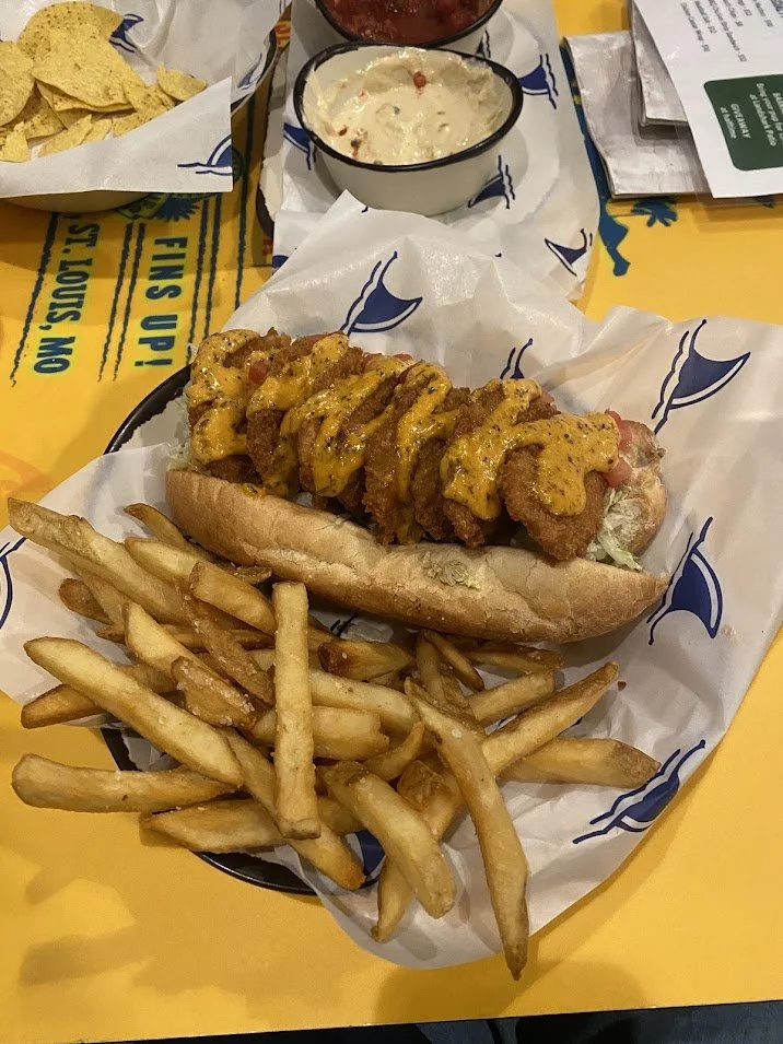Fried fish sandwich with French fries, served with dipping sauces and a side of tortilla chips.