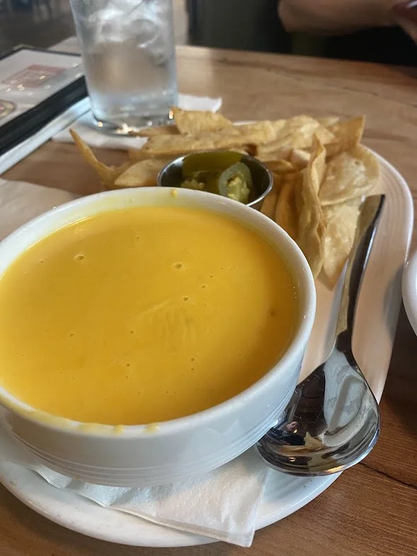 A bowl of yellow soup, possibly tomato or cheese soup, on a white plate with a spoon. In the background, there are fried yuca or plantain chips with sliced jalapeños, a glass of water with ice, and a person partially visible in a restaurant setting.