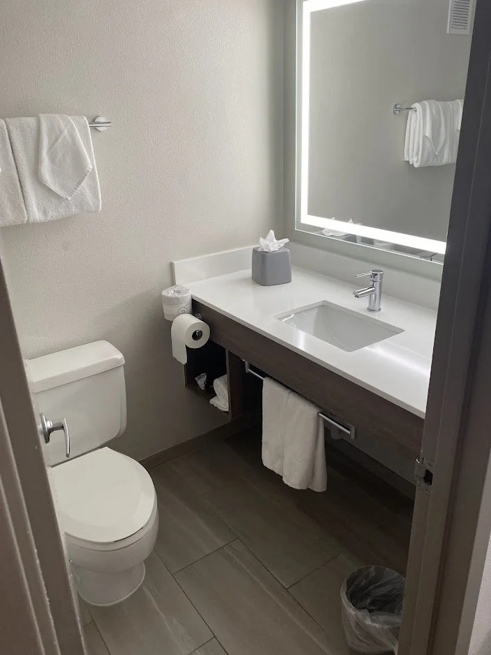A small, modern bathroom featuring a toilet, a floating vanity with a white countertop, a rectangular sink, a faucet, a large mirror with LED lighting, a towel bar with a white towel, a tissue box, and a small trash can.