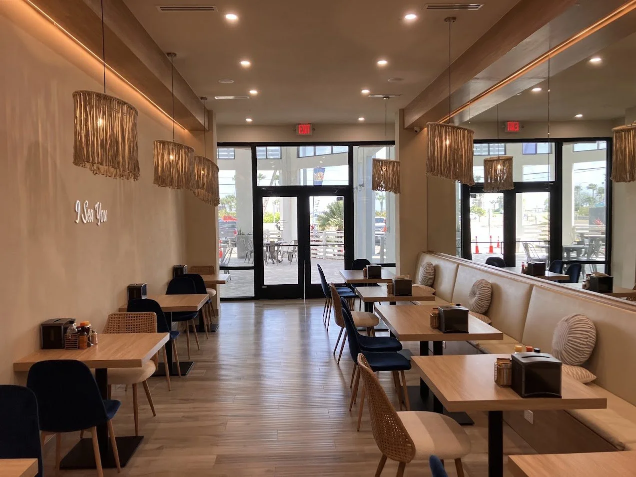 Empty restaurant with wooden floors, beige walls, and large windows. Small tables with chairs and cushions are arranged along one wall. Hanging light fixtures with fringed shades illuminate the space. Outside are parked cars and palm trees.