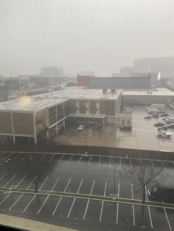 A cityscape view through a window shows an overcast, foggy day with a parking lot in the foreground, mostly empty with a few trees and parked cars. Behind, there is a large commercial building with a flat roof and multiple vehicles parked around it.