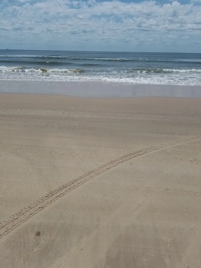 Empty sandy beach with tire tracks and the ocean in the background under a partly cloudy sky.