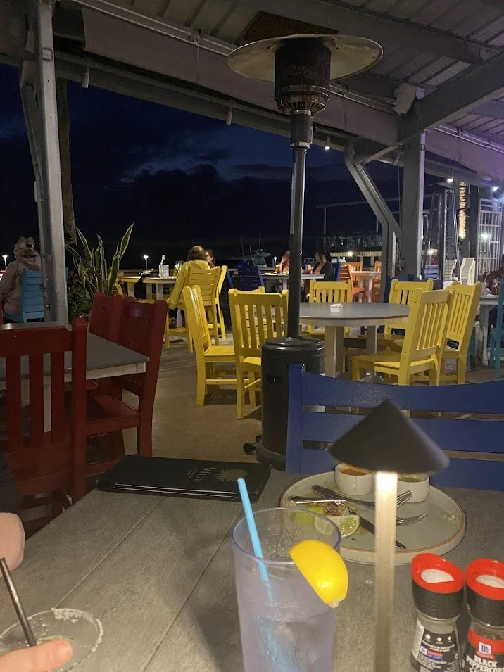 Nighttime outdoor restaurant with colorful tables and chairs, including yellow and blue chairs, a patio heater, and a drink with a lemon wedge on the table in the foreground.